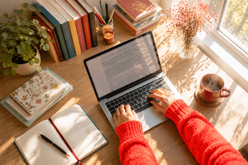 Hands typing on a laptop at a sunlit desk beside a window, with a steaming coffee, notebooks and colorful books.